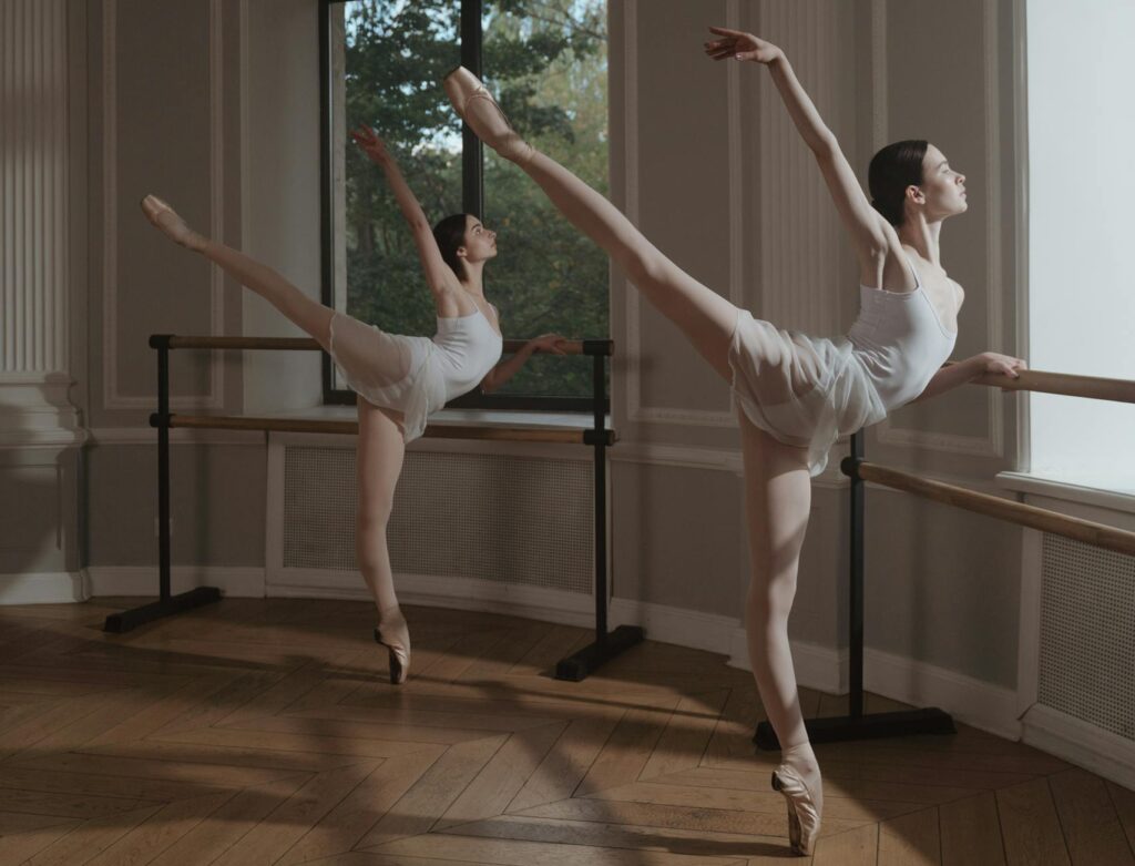 Two Ballerinas Doing Ballet Moves Beside Ballet Bar
